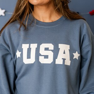 Puede incluir: Una sudadera azul con la palabra "USA" impresa en blanco en la parte delantera. La sudadera tiene una estrella blanca a cada lado de la palabra.
