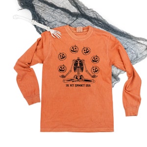 May include: Orange long-sleeve shirt with a black graphic of a skeleton meditating surrounded by jack-o'-lanterns. The text "IN MY SPOOKY ERA" is below the skeleton. A white skeleton hand and black fabric are in the background.