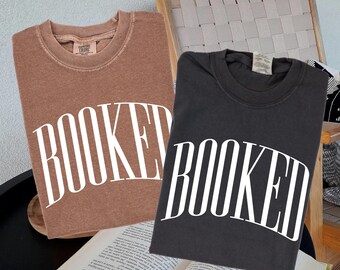 May include: Two crew neck sweatshirts, one brown and one black, with the word "BOOKED" in large white letters. An open book is in the foreground. The sweatshirts are on a black surface.