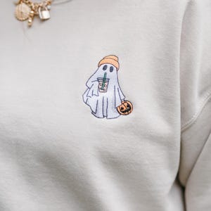 May include: Cream-colored sweatshirt with an embroidered Halloween ghost design. The ghost wears an orange beanie and holds a pumpkin and a beverage. The design is in black, white, and orange.