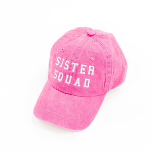 May include: Pink baseball cap with the text "Sister Squad" embroidered in white.