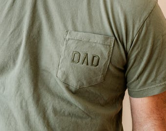 May include: Olive green t-shirt with a chest pocket. The pocket features the embroidered word "DAD" in a matching olive green thread. The shirt has short sleeves and a crew neck.