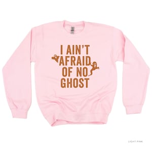 May include: Light pink crewneck sweatshirt with the phrase "I Ain't Afraid of No Ghost" in brown lettering. Two ghost graphics are on the sweatshirt. The sweatshirt is a solid light pink color.