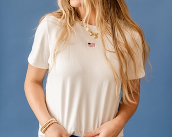 May include: Cream-colored t-shirt with a small embroidered American flag on the chest. The shirt is paired with blue bottoms and gold jewelry. The model is smiling, with long blonde hair, against a blue background.