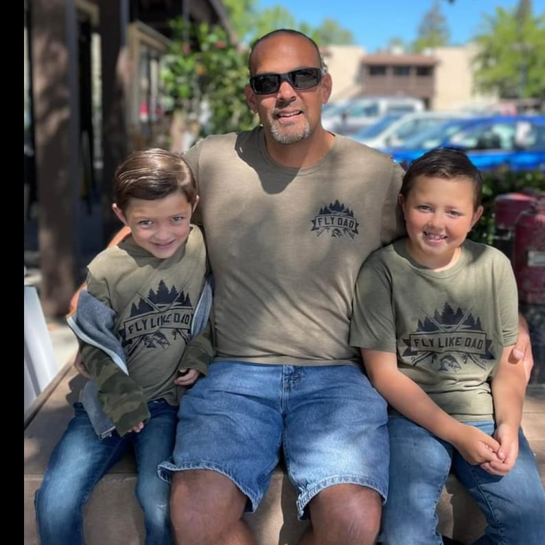 Op de afbeelding: Een vader en twee zonen dragen bijpassende groene T-shirts met een zwarte grafische print van een bergketen en de tekst "Fly Like Dad". De vader draagt een groen T-shirt met de tekst "Fly Dad".