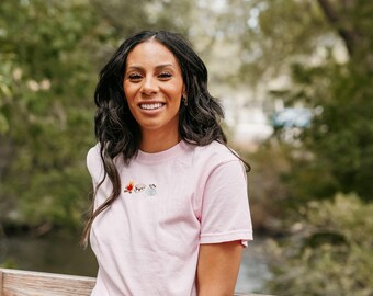 May include: A light pink t-shirt with a small embroidered design of a campfire, a marshmallow, and a mug. The shirt has short sleeves and is made of a soft material. The person is smiling and has long, dark hair.