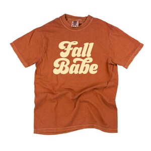 May include: A rust-colored t-shirt with the words "Fall Babe" in a cream-colored, retro font. The short-sleeved shirt has a crew neck and a slightly faded appearance, suggesting a vintage or worn-in style. The shirt is likely made of cotton or a similar fabric.