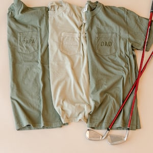 May include: Three olive green and tan t-shirts with pocket detail. The shirts have the words "PAPA", "GRANDPA", and "DAD" embroidered on the pockets. Two red golf clubs and two golf club heads are visible.