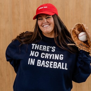 Puede incluir: Una mujer lleva una sudadera azul marino con el texto "THERE'S NO CRYING IN BASEBALL" en blanco. También lleva una gorra de béisbol roja que dice "GAME DAY" en blanco. Está sosteniendo un guante de béisbol de cuero marrón con una pelota de béisbol dentro.