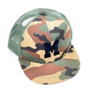 Könnte beinhalten: Eine Camouflage-Trucker-Kappe mit einem schwarzen Filzbuchstaben "M" auf der Vorderseite. Die Kappe hat ein Netzrückenpanel.