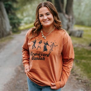 May include: An orange long-sleeve shirt with a graphic of dancing monsters and the text "It Was a Graveyard Smash". The shirt is made of a soft material.