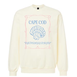 Op de afbeelding: Crèmekleurige sweatshirt met de tekst "CAPE COD" in blauw, een blauwe schelpgrafiek en een roze kader. Daaronder staat de tekst: "waar zoutgezoende briesjes dansen door verwelkte shingles en meeuwen kletsen over zandduinen."