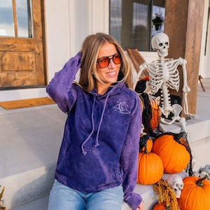 May include: A purple acid-wash hooded sweatshirt featuring a small white embroidered graphic on the left chest depicting a crescent moon, stars, and clouds. The long-sleeve pullover includes a front kangaroo pocket and matching purple drawstrings.
