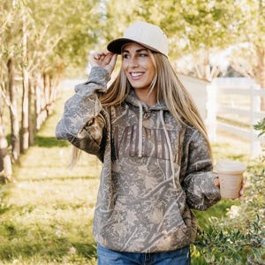 May include: A camouflage-patterned hoodie with the word "Youth" across the chest. The person is wearing a tan baseball cap with a brown brim and holding a coffee cup. The person is wearing blue jeans.