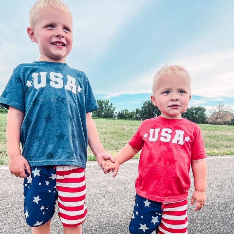May include: Two young boys wearing red and blue shirts with the word "USA" printed on them. They are both wearing matching red, white, and blue star-patterned swim trunks.