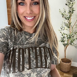 Op de afbeelding: Een camouflage-patroon T-shirt met het woord "MOTHER" in grote, bruine blokletters. Het shirt heeft korte mouwen en een ronde hals. Op de achtergrond een decoratieve plant in een gevlochten mand.