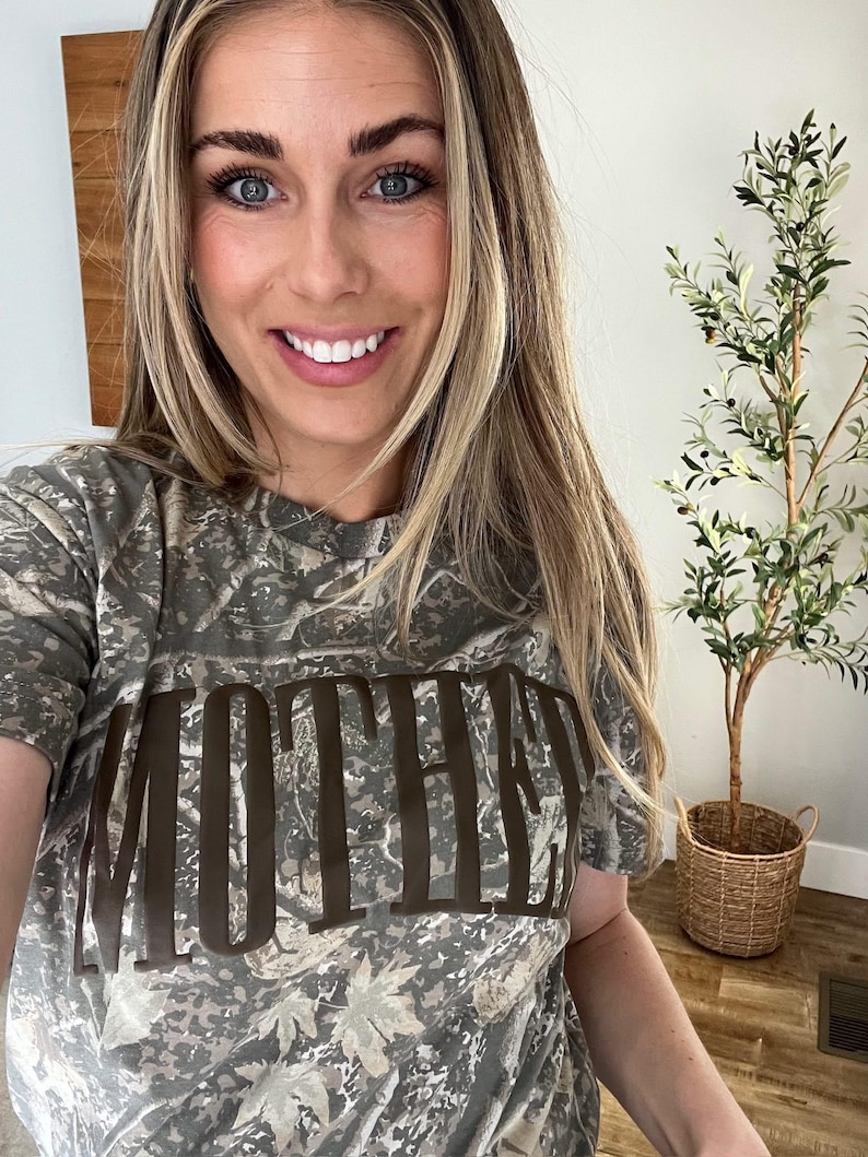 Op de afbeelding: Een camouflage-patroon T-shirt met het woord "MOTHER" in grote, bruine blokletters. Het shirt heeft korte mouwen en een ronde hals. Op de achtergrond een decoratieve plant in een gevlochten mand.