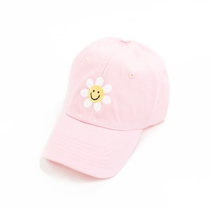 May include: A pink baseball cap with a white daisy embroidered on the front. The daisy has a yellow smiley face in the center.
