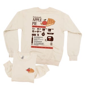 Op de afbeelding: Crèmekleurig sweatshirt met lange mouwen en shirt met korte mouwen. De achterkant van het sweatshirt heeft een recept voor zelfgemaakte appeltaart met illustraties. De voorkant van het shirt heeft een kleine appeltaartafbeelding. Tekst bevat "homemade APPLE PIE".