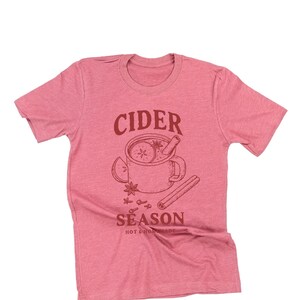 May include: A pink t-shirt with a crew neck. The shirt features a brown graphic of a mug of cider with cinnamon sticks, star anise, and orange slices. The words "CIDER SEASON" are printed above and below the mug.