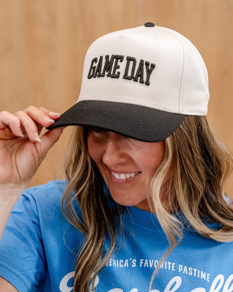 May include: A white and black baseball cap with the text "GAME DAY" in black embroidered letters.