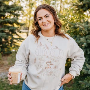 May include: A beige sweatshirt with a fall-themed design featuring pumpkins, leaves, and cinnamon sticks. The text "pick of the patch" is printed on the front. The person is holding a coffee cup.