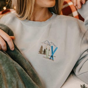May include: A light blue sweatshirt with an embroidered design of mountains, skis, and trees. The design features blue skis, green and brown trees, and gray mountains. The sweatshirt is a crew neck style.