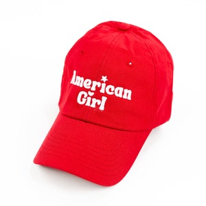 Op de afbeelding: Rode baseballpet met witte geborduurde tekst die "American Girl" luidt met een kleine ster boven het woord "Girl".