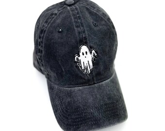 May include: A black, distressed baseball cap with a white embroidered ghost design on the front. The cap has a curved brim and a fabric-covered button on top. The hat is made of a soft, worn material.