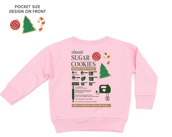 Può includere: Felpa rosa chiaro con una grafica della ricetta dei biscotti di zucchero sul retro. La ricetta include illustrazioni di ingredienti e istruzioni. La parte anteriore presenta un design tascabile con un ornamento natalizio, un albero e un bastoncino di zucchero.