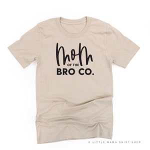 May include: A tan crew neck t-shirt with the text "Mom of the Bro Co." in black script. The shirt is made of a soft material and is likely for casual wear. The text is centered on the front of the shirt.