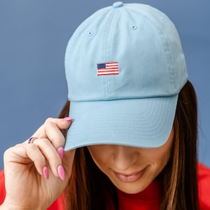 May include: Light blue baseball cap with a small embroidered American flag on the front. The cap is made of a soft material and has a curved brim. The cap is a classic style and is suitable for everyday wear.