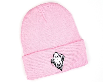 Puede incluir: Un gorro rosa claro con un borde doblado. El gorro presenta un diseño de fantasma bordado en blanco y gris en la parte delantera. El gorro está hecho de un material suave y tejido.