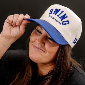 May include: A white baseball cap with a blue brim and the text "SWING BATTER BATTER" in blue embroidery. The number "13" is embroidered in blue on the right side of the cap.