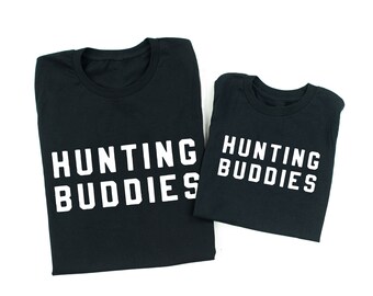 May include: Two black t-shirts with white text that reads "Hunting Buddies". The larger shirt has the text "Hunting Buddies" on the front, while the smaller shirt has the text "Hunting Buddies" on the back.