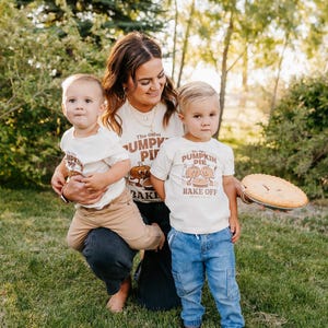 May include: A family poses outdoors, wearing matching cream-colored t-shirts with a pumpkin pie graphic and the text "The Great Pumpkin Pie Bake Off." One person holds a pie, and the others are on the grass.