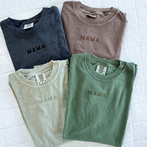 May include: Four t-shirts in different colors, each with the word "MAMA" embroidered on the front. The shirts are in black, brown, green, and light green.