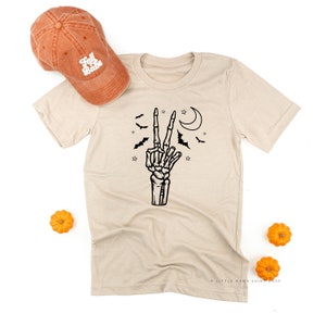Peut inclure: T-shirt beige avec une main de squelette noire faisant le signe de la paix, entourée de chauves-souris, d'étoiles et d'un croissant de lune. Une casquette de baseball orange avec le texte "Fall Babe" est à côté du t-shirt. De petites citrouilles oranges sont également dans l'image.