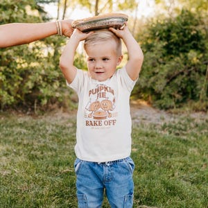 May include: A young child wearing a cream-colored t-shirt with a pumpkin pie graphic and the words "Pumpkin Pie Bake Off." The child is holding a pie tin on their head. They are wearing blue denim jeans and standing on green grass.