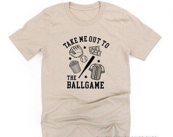 Peut inclure: Un t-shirt beige à manches courtes avec des graphismes noirs imprimés représentant un gant de baseball, une balle, une batte, du pop-corn et des billets. Le texte "TAKE ME OUT TO THE BALLGAME" est écrit dans une police athlétique.
