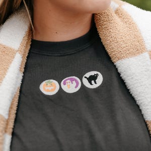 Puede incluir: Sudadera gris oscuro con tres parches bordados con temática de Halloween: una calabaza, una bruja y un gato negro. La persona lleva una chaqueta a cuadros beige y blanca.
