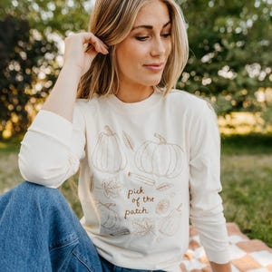May include: Cream-colored long-sleeve shirt with a crew neck. The shirt features brown line drawings of pumpkins, leaves, and the text "pick of the patch". The shirt is worn by a person sitting outside on a blanket.