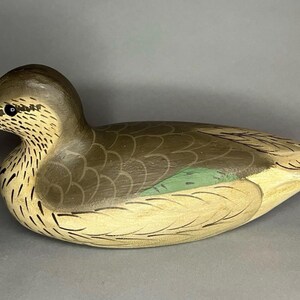 May include: A hand-painted wooden duck figurine. The duck features a detailed design with a brown head, back, and wings, accented with green and cream-colored details. The beak is yellow. This decorative item is likely for home decor.
