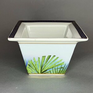 May include: A square, white ceramic bowl with a blue and gold geometric border. The bowl features a light blue background with green palm fronds. A gold band accents the base of the bowl.