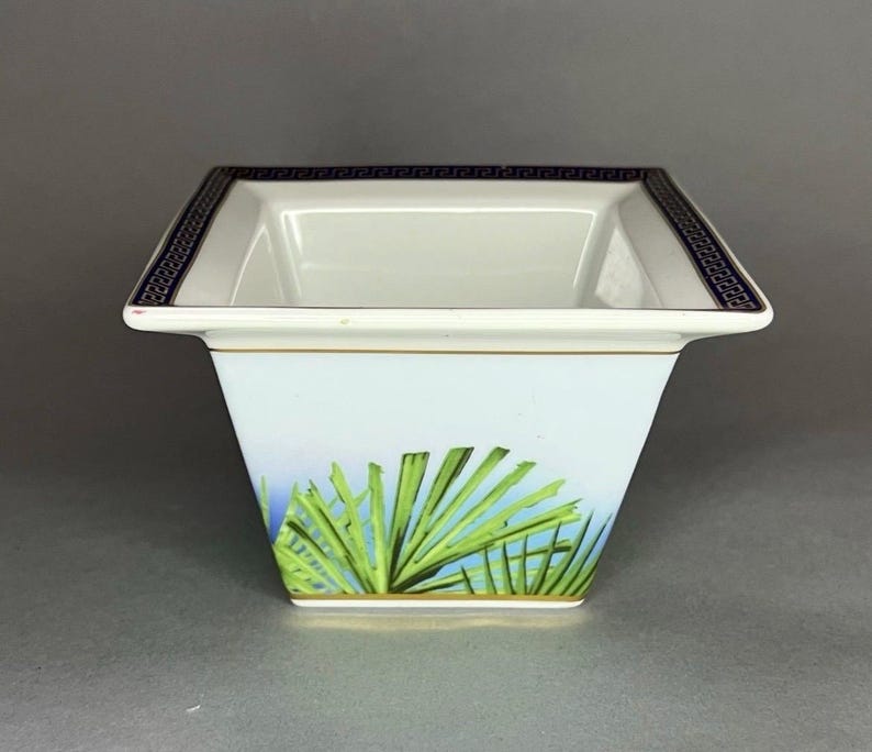 May include: A square, white ceramic bowl with a blue and gold geometric border. The bowl features a light blue background with green palm fronds. A gold band accents the base of the bowl.