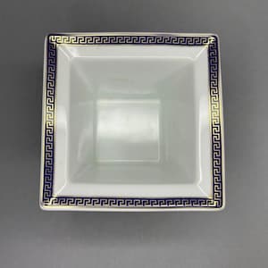 May include: A square, white ceramic bowl with a blue and gold Greek key border design. The bowl has a slightly flared shape and a smooth, glossy finish. The interior is plain white.