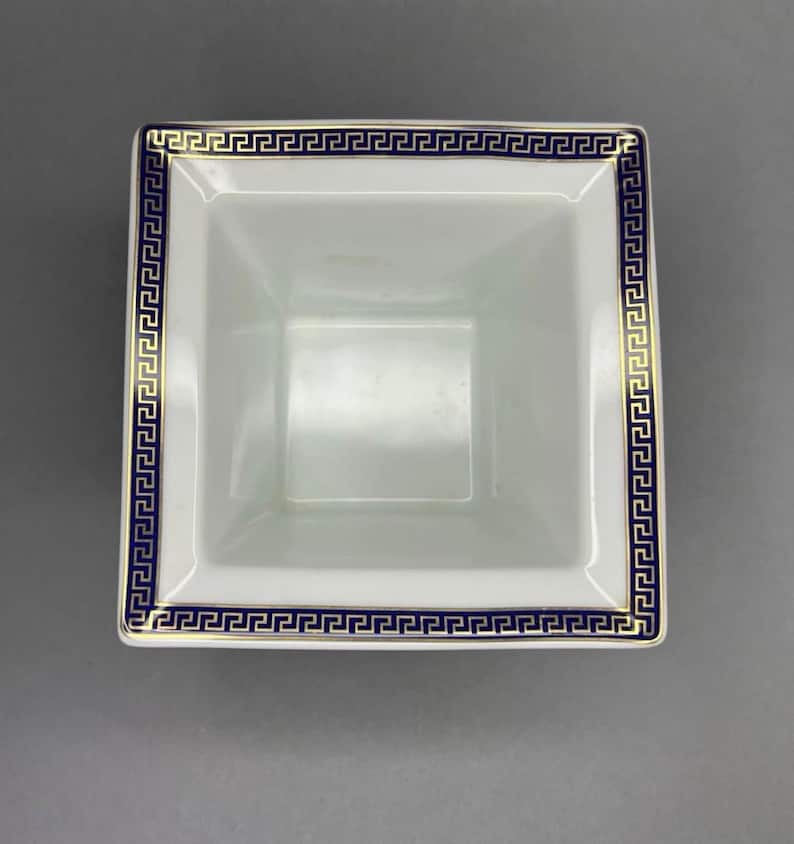 May include: A square, white ceramic bowl with a blue and gold Greek key border design. The bowl has a slightly flared shape and a smooth, glossy finish. The interior is plain white.