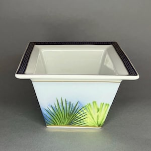 May include: A square, white porcelain bowl with a light blue and green palm leaf design. The rim features a dark blue Greek key pattern and a gold trim. The bowl has a slightly flared shape.