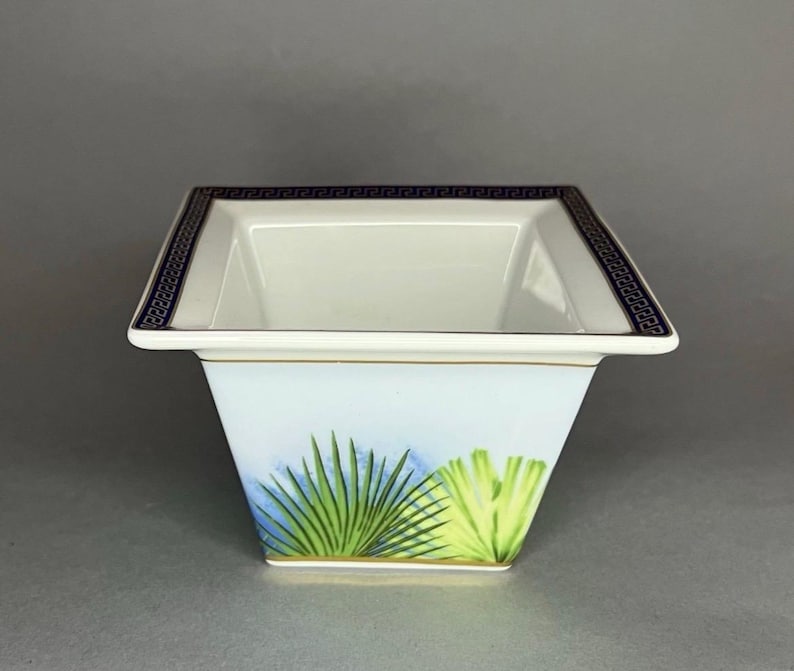 May include: A square, white porcelain bowl with a light blue and green palm leaf design. The rim features a dark blue Greek key pattern and a gold trim. The bowl has a slightly flared shape.
