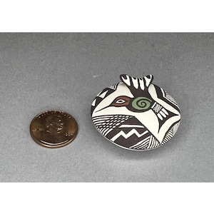 May include: A small, round, hand-painted ceramic vessel with a unique design. The vessel features a black and white geometric pattern, with accents of green and brown. A penny is next to the vessel for size comparison.